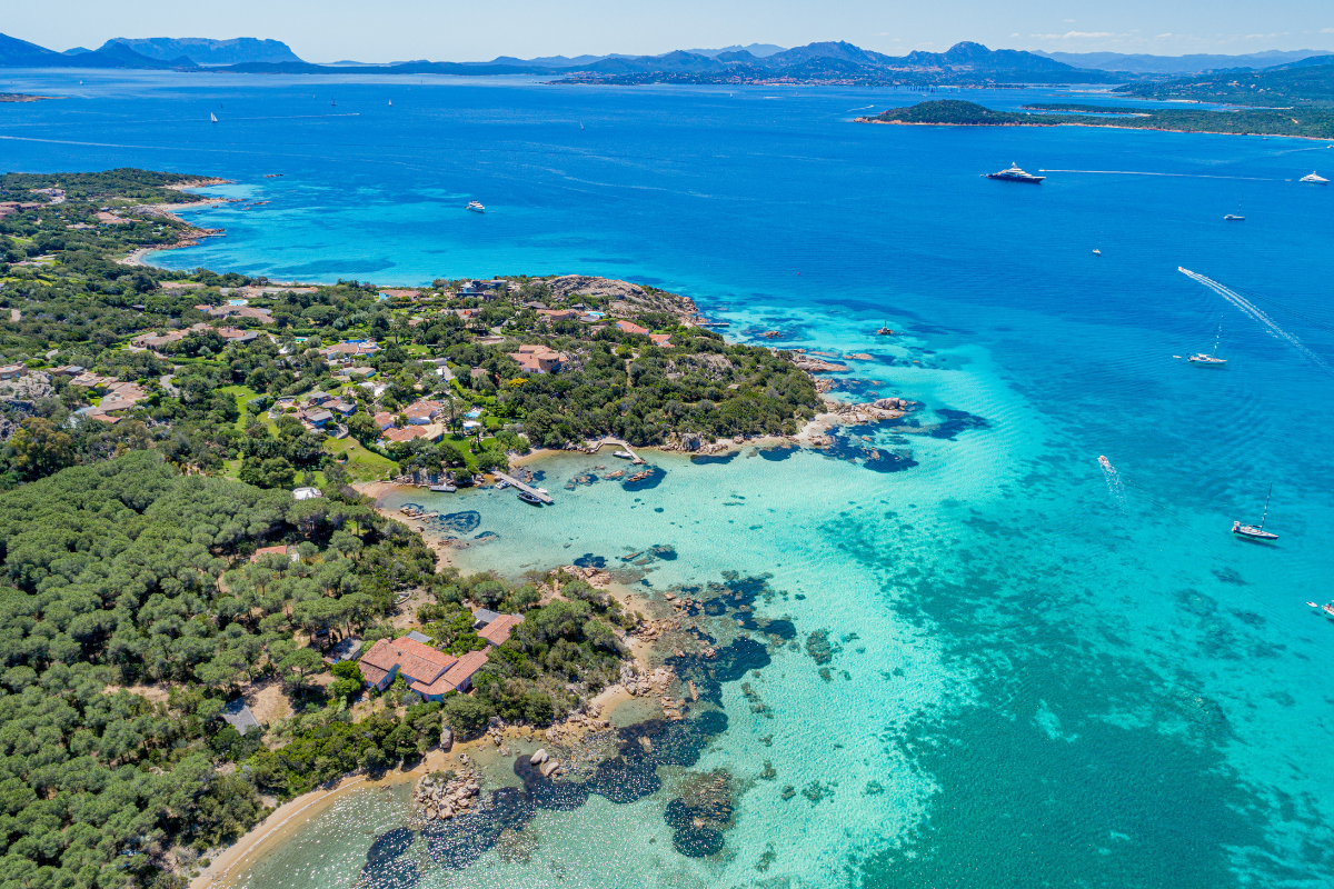 Costa Smeralda, Sardinija – jachta plaukia žaliai žydru vandeniu, pušynai ir kalvos pakrantėje