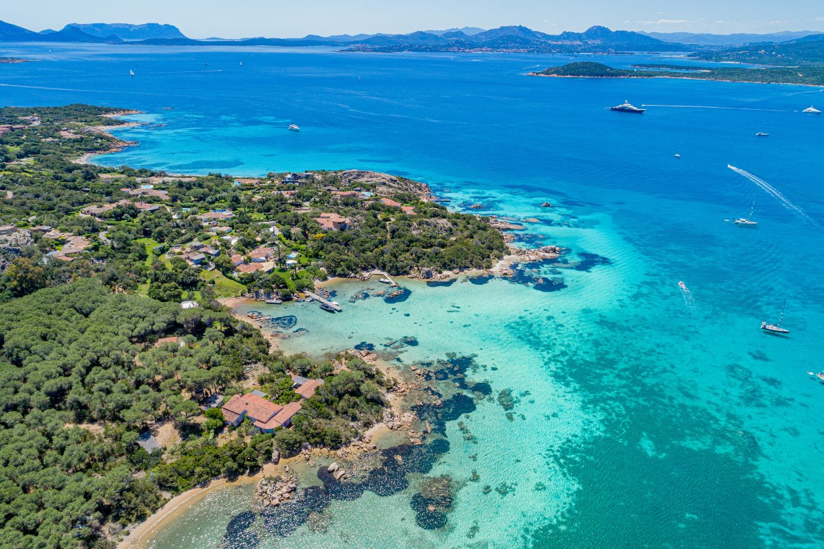 Costa Smeralda, Sardinija – jachta plaukia žaliai žydru vandeniu, pušynai ir kalvos pakrantėje